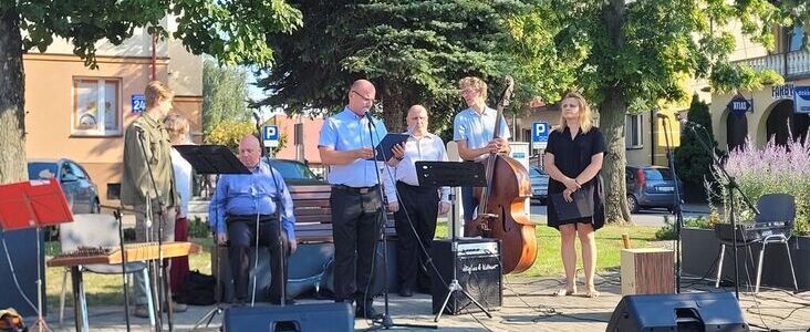 Grupa ludzi uczestniczy w wydarzeniu na zewnątrz, stoi na przemian przy mikrofonie, z instrumentami muzycznymi, w tym kontrabasem. Po lewej widoczni są siedzący widzowie.