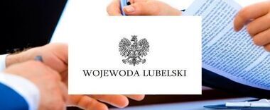 Osoba trzymająca dokument z godłem Polski i napisem "WOJEWODA LUBELSKI". Częściowo widoczne długopisy i papier na biurku.