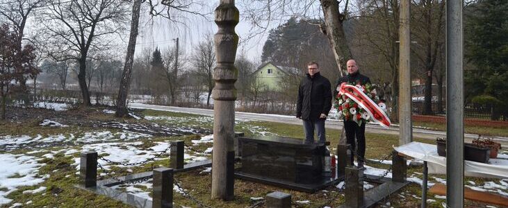 Mężczyźni przy grobie z kamiennym krzyżem na tle zimowego krajobrazu. Jeden trzyma wieniec w barwach narodowych Polski. Scena rozgrywa się przy niewielkiej, zaśnieżonej drodze.