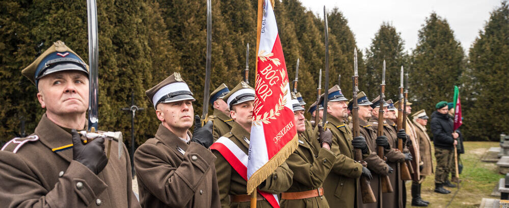 Grupa mężczyzn w historycznych mundurach stoi w szeregu obok sztandaru, uczestnicząc w uroczystości na zewnątrz. Tło stanowią wysokie, zielone drzewa.