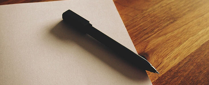 Na zdjęciu widoczny jest czarny długopis leżący ukośnie na pustej kartce papieru. Całość znajduje się na drewnianym stole. Oświetlenie jest naturalne, co tworzy ciepłą atmosferę.