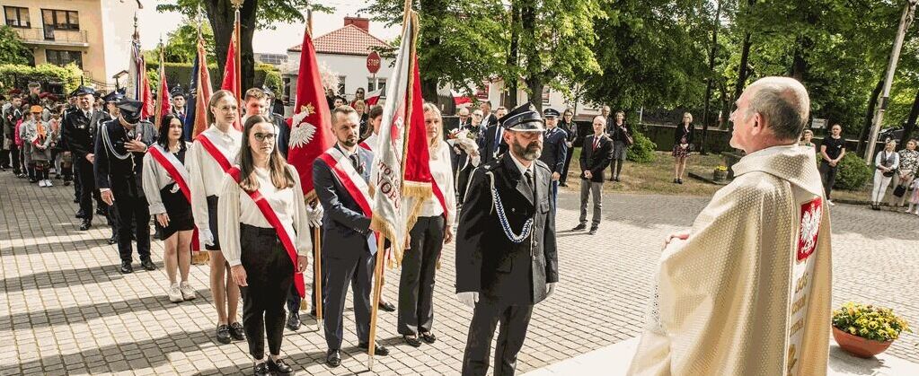 Grupa ludzi w oficjalnych strojach ustawia się w szeregu na zewnątrz. Niektórzy trzymają polskie flagi. Osoba w sutannie stoi naprzeciwko grupy. Scena odbywa się w parku.