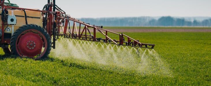 Ciągnik wyposażony w opryskiwacz rozpyla pestycydy na polu uprawnym, pokrytym zieloną trawą. W tle widoczna jest linia drzew na horyzoncie pod błękitnym niebem.