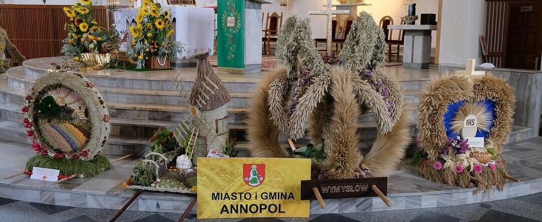 Wnętrze kościoła z dekoracjami dożynkowymi: wieńce z kłosów zboża, kwiaty słoneczników i symbole religijne. Na przodzie tablica z napisem "Miasto i Gmina Annopol".