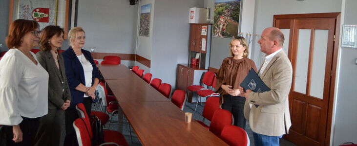 Na zdjęciu widać grupę osób stojących w sali konferencyjnej z czerwonymi krzesłami i długim stołem. Jedna osoba trzyma teczkę. W tle widać zegar na ścianie i różne dekoracje.