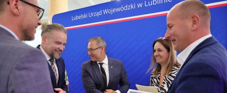 Grupa osób w eleganckich strojach rozmawia przed niebieskim tłem z napisem "Lubelski Urząd Wojewódzki w Lublinie." Jedna osoba trzyma certyfikat lub dyplom. Wszyscy wyglądają na zadowolonych.