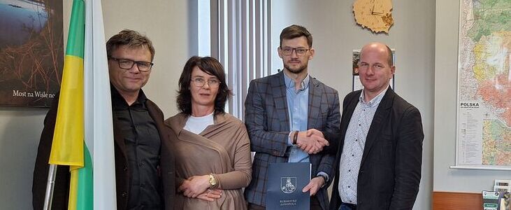 Czworo ludzi stoi w biurze obok flagi i mapy na ścianie. Dwie osoby trzymają dokument z herbem. W tle widoczne są przedmioty biurowe i zegar wskazujący godzinę 12:17. Po lewej stronie jest plakat z mostem.