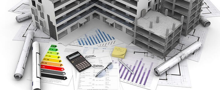 Widok z góry na biurko architekta z makietami budynków, planami budowlanymi, wykresami, kalkulatorem, długopisem i kolorowymi dokumentami. W tle zwinięte plany architektoniczne.