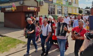 Zdjęcie przedstawia grupę ludzi uczestniczących w ulicznym wydarzeniu, z mężczyznami grającymi na akordeonie i gitarze. Widoczny budynek z reklamami i niebo z chmurami.