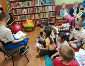 Zdjęcie przedstawia dzieci skupione na kanapie i dywanie. Na pierwszym planie jest chłopiec siedzący na krześle i czytający książkę. W tle stoją regały z książkami.