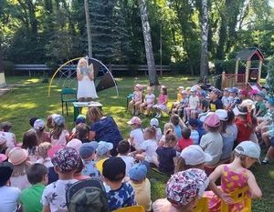 Na zielonej trawie na żółtych krzesełkach siedzą przedszkolaki, dalej stoi bibliotekarz z mikrofonem i z książką w ręku, w tle znajdują się drzewa i plac zabaw