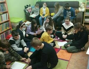 Zdjęcie przedstawia grupę dzieci. Część z nich siedzi na kolorowym dywanie, a część na brązowej kanapie. Po lewej stronie widoczny jest kawałek regału z książkami. Dzieci rozwiązują zagadki.
