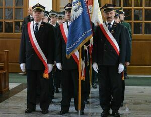 Mężczyźni w mundurach z szarfami stoją w szeregu, trzymając flagę z orłem i napisem, w tle drzwi i okna w eleganckim wnętrzu.