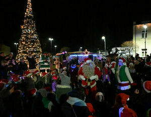 Grupa ludzi zgromadzona na wieczornym świątecznym wydarzeniu na zewnątrz, z choinką, Świętym Mikołajem i elfem w tle.