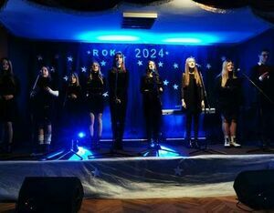 Grupa ludzi w czarnym ubraniu stoi na scenie z mikrofonami, w tle napis "ROK 2024" i gwiazdy, wyglądają na zespół podczas występu.