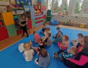 Na zdjęciu widać grupę dzieci siedzących na niebieskim dywanie. W tle widnieją okna ,na szybach są naklejone motyle. Przed dziećmi siedzi na krześle bibliotekarka czytająca książkę. Za nią ustawiony jest kolorowy regał z zabawkami.