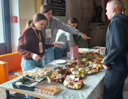 Osoby stoją przy stole z różnorodnymi potrawami podczas wydarzenia, wybierają jedzenie przy asyście wolontariusza.