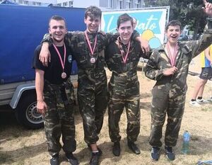 Czterej uśmiechnięci mężczyźni w kamuflażowych mundurach stoją obok siebie z medalami na szyjach, celebrują sukces, w tle niewyraźny pojazd i baner.
