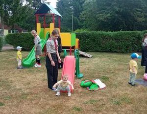 Opis zdjęcia: Plac zabaw z dziećmi i opiekunem. Dziecko schodzi po zjeżdżalni, inne bawią się w tle. Jest trawa i drzewa wokół.