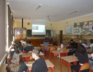 Zdjęcie przedstawia uczniów siedzących w klasie szkolnej, skupionych na prezentacji, którą prowadzi osoba stojąca przy tablicy wyposażonej w projektor.