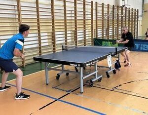 Dwóch mężczyzn gra w tenisa stołowego w sali gimnastycznej. Jeden z nich jest w trakcie wykonania serwisu.