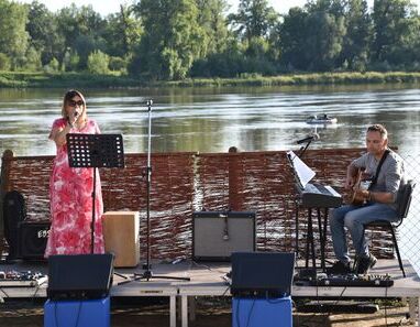 Kobieta śpiewająca i mężczyzna grający na gitarze na scenie na tle rzeki.