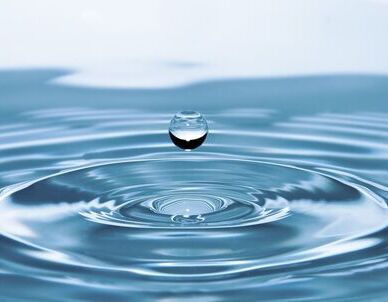 Jasnoniebieska woda z idealnie okrągłą kroplą unoszącą się tuż nad powierzchnią, tworząc koncentryczne fale.