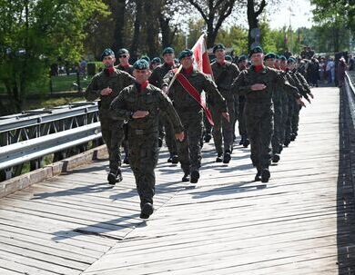 Grupa żołnierzy w mundurach maszeruje po drewnianym moście, jeden z nich trzyma flagę. Na tle drzew i przechodniów.