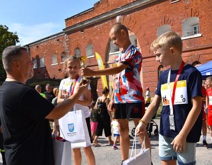 Na zdjęciu jest grupa dzieci odbierająca nagrody na zewnątrz. Dziecko w środku trzyma trofeum, a dorosły wręcza prezenty. W tle widoczna ceglana ściana.