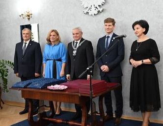 Grupa pięciu osób stoi przy drewnianym biurku z mikrofonem w eleganckim pomieszczeniu z herbem narodowym na ścianie.