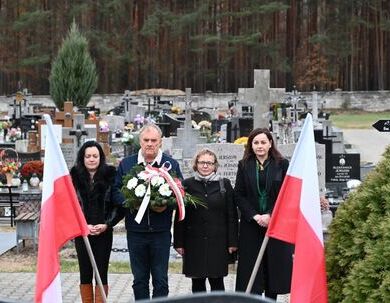 Grupa czterech osób stojących na cmentarzu z wieńcem, otoczona flagami Polski, w tle mogiły i drzewa.