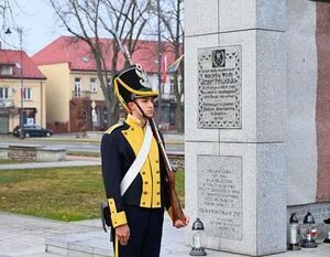 Straż honorowa w mundurze z epoki, stoi obok kamiennego pomnika z tablicą pamiątkową, w tle widoczne są drzewa i budynki.