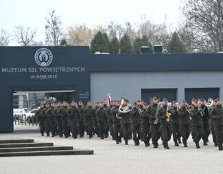 Orkiestra wojskowa maszeruje przed muzeum, ubrana w mundury maskujące, z instrumentami dętymi i perkusyjnymi.