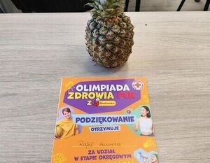 Ananas z czerwonym sercem na czubku stoi na drewnianym stole. Obok niego kolorowa ulotka informuje o Olimpiadzie Zdrowia PCK, zawiera podziękowania i ilustracje dzieci. W tle widać krzesła.
