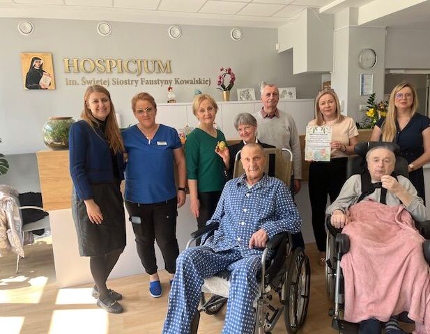 Grupa ludzi w hospicjum pozuje do zdjęcia. Dwie osoby siedzą na wózkach inwalidzkich, otoczone przez uśmiechnięty personel. W tle widoczny jest napis informujący o nazwie placówki.