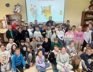 Grupa dzieci i kilku dorosłych pozuje w sali z projekcją na ścianie. Na ekranie widać postacie z animacji. W tle znajdują się półki z książkami i dekoracje.