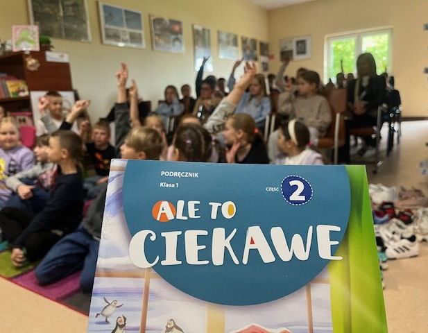 Podręcznik "Ale to ciekawe" trzymany na pierwszym planie. W tle dzieci siedzą w klasie, niektóre z rękami podniesionymi do góry. Pomieszczenie jest jasne, z dużymi oknami.