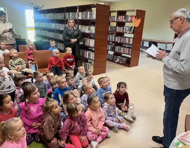 Starszy mężczyzna czyta książkę dzieciom siedzącym na podłodze w bibliotece. Dookoła półki z książkami, dzieci ubrane są kolorowo i patrzą z zainteresowaniem.