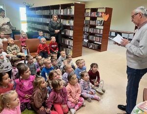 Starszy mężczyzna czyta książkę dzieciom siedzącym na podłodze w bibliotece. Dookoła półki z książkami, dzieci ubrane są kolorowo i patrzą z zainteresowaniem.