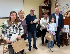 Grupa sześciu osób stoi w pomieszczeniu. Dwie z nich trzymają dyplomy i torby papierowe. W tle plakat i półki z książkami oraz maskotkami. Na ścianie ekran projekcyjny. Wszyscy są uśmiechnięci.