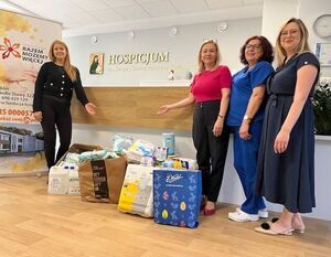 Cztery kobiety stoją w holu hospicjum obok darów, takich jak pieluchy i środki higieniczne. Na ścianie widać napis "Hospicjum". Każda z kobiet jest elegancko ubrana i uśmiechnięta.