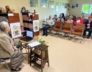 Spotkanie dla seniorów w bibliotece. Kobieta prezentuje materiał na laptopie. Grupa osób siedzi na krzesłach, słuchając z zainteresowaniem. W tle półki z książkami i plakaty. Na ścianie ozdoby i zdjęcia.