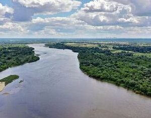 Rzeka z płynącą wodą otoczona bujną zielenią i roślinnością. W tle widać rozległe pola oraz miasto na horyzoncie. Niebo jest częściowo zachmurzone, co tworzy malowniczy krajobraz.