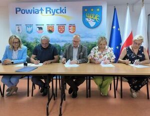 Pięć osób siedzi przy stole, podpisując dokumenty. Za nimi widoczna jest tablica z nazwami gmin i herbami, a także flagi Polski i Unii Europejskiej. W tle znajduje się plakat z napisem "Powiat Rycki".