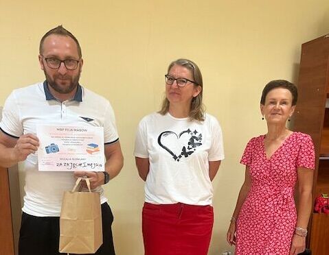 Trzy osoby stoją w pomieszczeniu. Mężczyzna trzyma dyplom i torbę, obok niego dwie kobiety. Kobieta w środku ma białą koszulkę i czerwoną spódnicę, a kobieta po prawej ma czerwoną sukienkę. W tle jasna ściana.