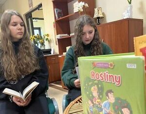 Dwie dziewczyny siedzą na krzesłach w pomieszczeniu z książkami. Jedna z nich trzyma otwartą książkę, a druga patrzy w dół. Na pierwszym planie leży książka o roślinach na stoliku.