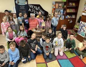 Grupa dzieci i dorosła osoba siedzą w bibliotece na kolorowym dywanie. Dookoła widać książki na półkach oraz plakat promujący spotkanie autorskie. Dzieci większość patrzy w stronę aparatu.