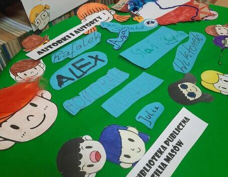 Kolorowe, ręcznie wykonane ozdoby, napisy i postacie z papieru leżą na zielonym tle. W tle widać półki z książkami. Na przyczepionej kartce napis: "ANIMACJA DO WIERSZA... KTO TY JESTEŚ?" oraz informacje o bibliotece.