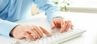 Osoba w niebieskiej koszuli pracuje przy biurku, używając białej klawiatury komputera.