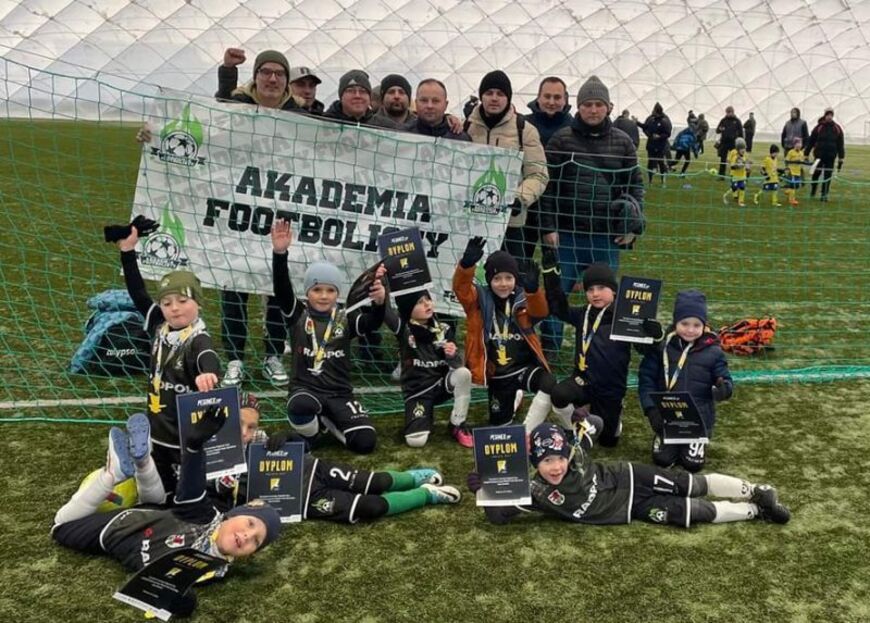 Grupa dzieci w czarnych sportowych strojach i medale, z trenerami i banerem "Akademia Football'u", na tle boiska i innej drużyny.